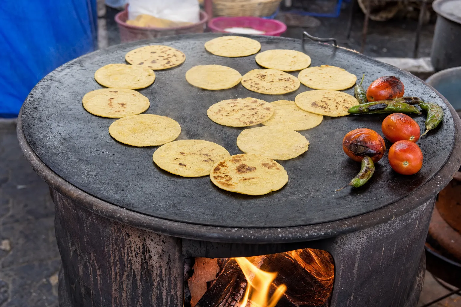 薪の火にかけた、メキシコの市場の大きな丸い鉄のコマル。小さな黄色いコーントルティーヤがいくつも並んで焼け、右端で黒く焦げたトマトと青唐辛子を焼いている