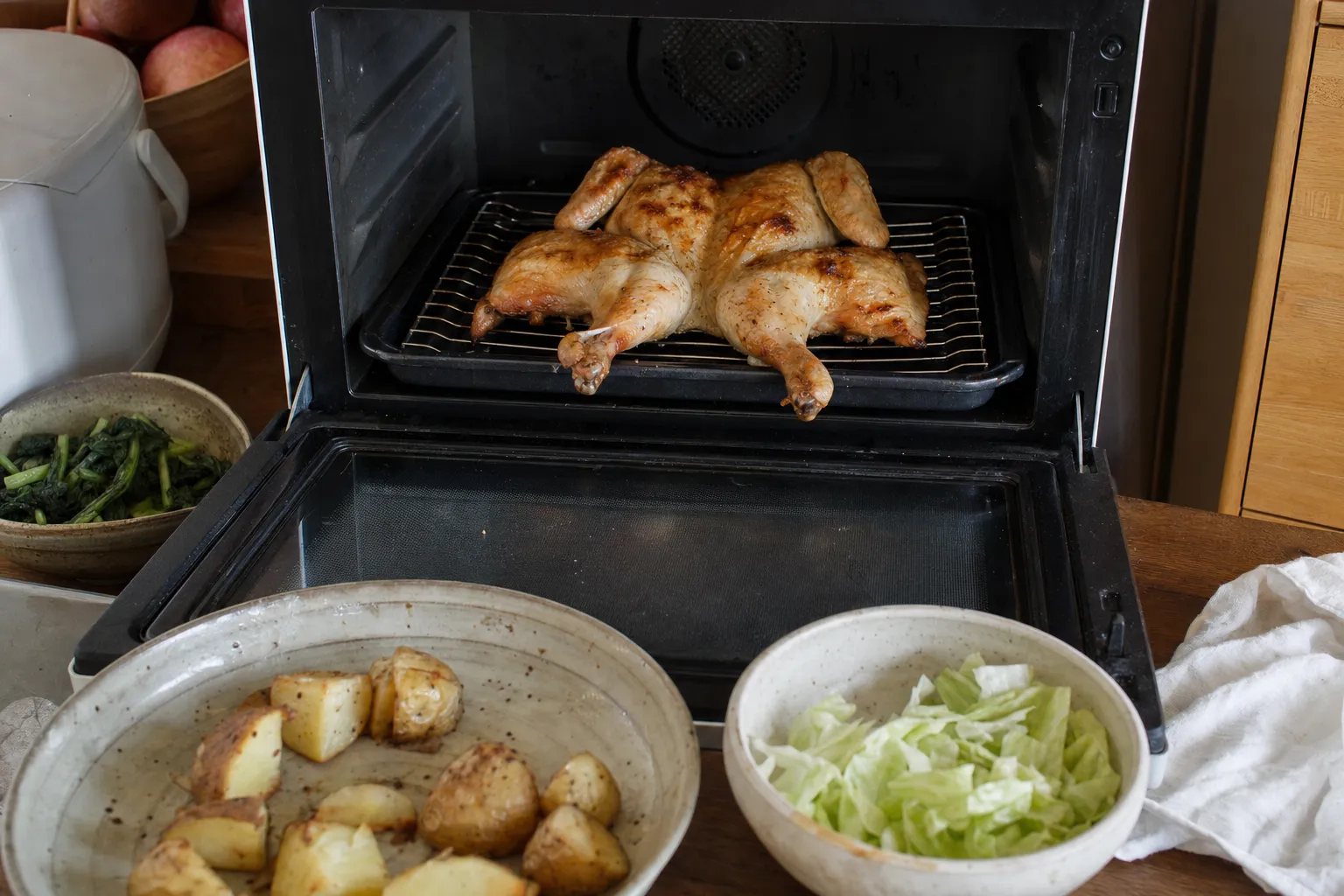 扉を開けた家庭のオーブンレンジの中で、角皿にのせた焼き網の上に置いた背開きの丸鶏。皮は半分ほどきつね色に焼けている