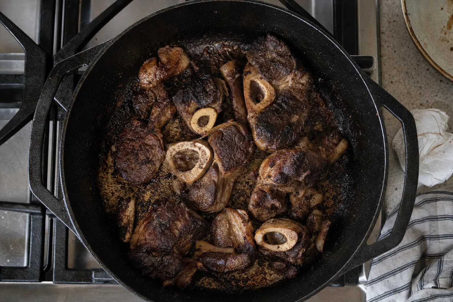 コンロにかけた黒い鋳鉄のダッチオーブンを上から見たところ。輪切りの牛すね肉の全面に濃い焼き色がつき、鍋底に茶色い焼きつきが残っている