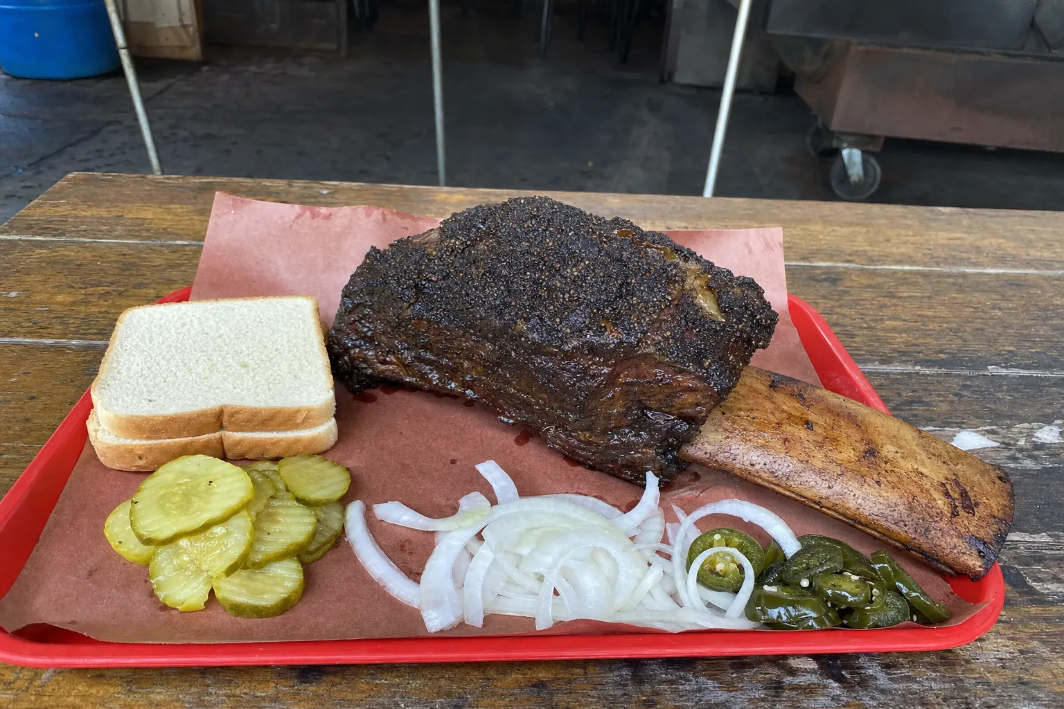 赤いトレーにピンクの紙を敷いてのせた、長い骨が1本ついた大きなビーフリブ。食パン、ピクルス、玉ねぎの薄切り、ハラペーニョの酢漬けを添えて