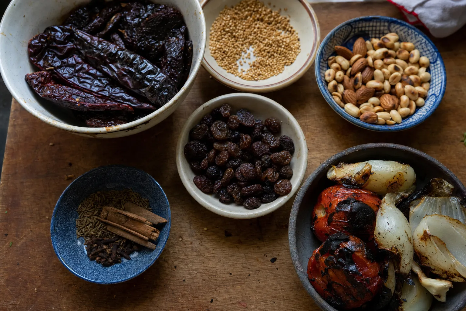 木のまな板に並んだ、焼いたモレの材料の器。揚げて黒っぽくなった乾燥唐辛子、炒ったごま、アーモンドとピーナッツ、揚げたレーズン、シナモンとクローブ、焦げ目のついたトマトと玉ねぎ